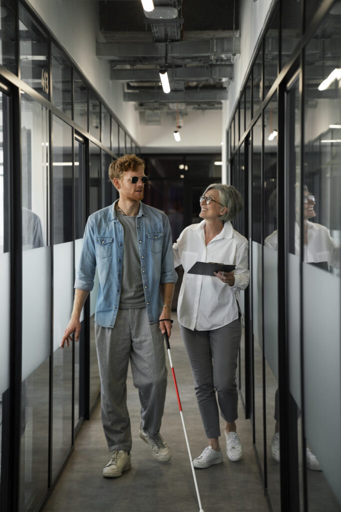 A man using a white cane walks down a modern office hallway with glass walls. He is wearing sunglasses and casual clothes. Next to him walks a woman with short grey hair, holding a clipboard and gently touching his shoulder as they smile at each other.